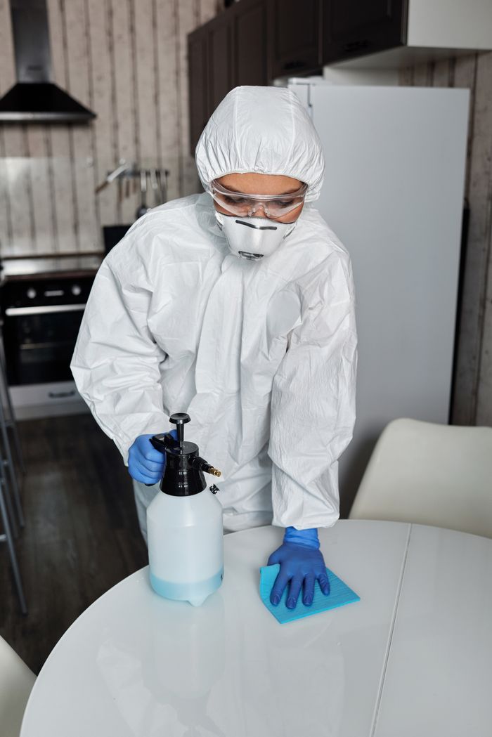 who-we-are A person in full PPE disinfecting a table indoors for hygiene and safety.